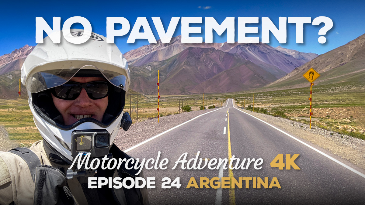 A man in a motorcycle helmet standing in front of an open road through the mountains.